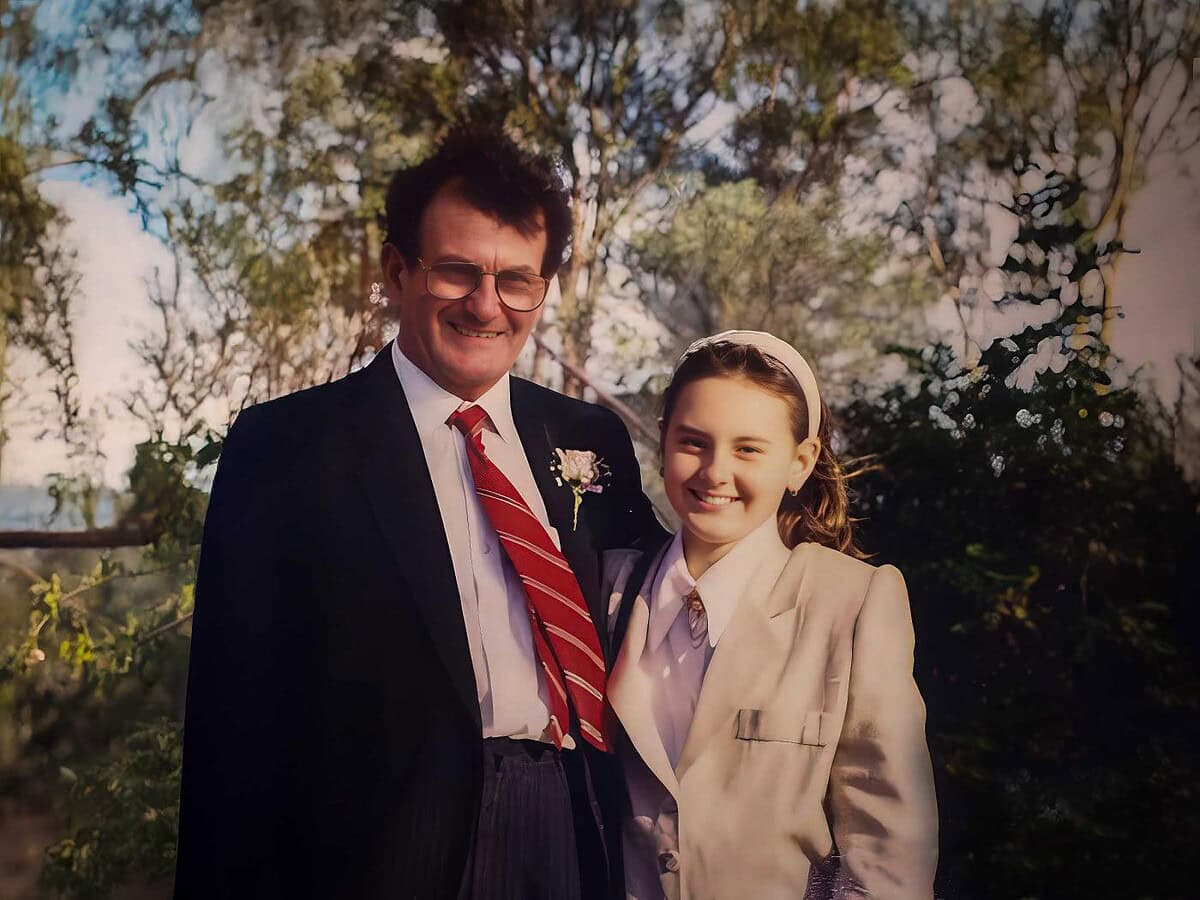 Enjoying a happy moment together outdoors, a man and a young girl smile warmly at the camera amidst lush greenery and blooming trees, capturing a joyful occasion or special event.